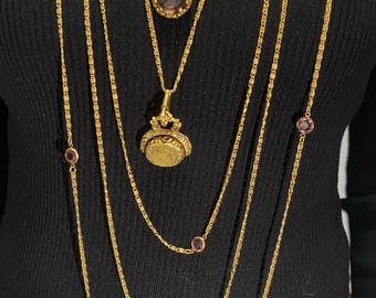 May include: Several gold necklaces of varying lengths, some with small, round, purple gemstones. One necklace features a pendant with a round, purple gemstone. Another has a coin-shaped pendant. The necklaces are layered against a black background.