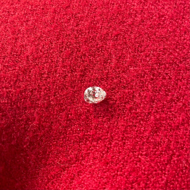 May include: A pear-shaped diamond, sparkling with facets, rests on a vibrant red textured surface. The gemstone's cut and clarity are highlighted against the contrasting background, showcasing its brilliance.