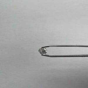 May include: A small, round, clear diamond held by a pair of silver tweezers. The diamond sparkles against a plain white background, showcasing its facets and brilliance. The tweezers are in sharp focus, highlighting the gem's clarity.