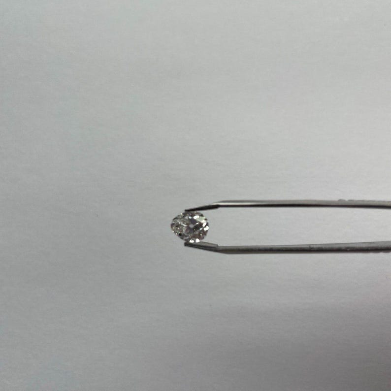 May include: A small, round, clear diamond held by a pair of silver tweezers. The diamond sparkles against a plain white background, showcasing its facets and brilliance. The tweezers are in sharp focus, highlighting the gem's clarity.
