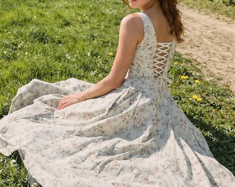 May include: A sleeveless floral print dress with a lace-up back detail. The dress is light-colored with a pattern of small pink flowers. The person is sitting on a grassy area with a path and trees in the background.