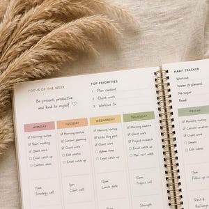 May include: An open spiral-bound weekly planner with cream-colored pages featuring sections for focus of the week, top priorities, a habit tracker, and daily schedules. The planner includes handwritten notes and a gold pen resting on the side.