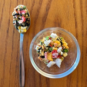 May include: A stainless steel spoon holds a portion of colorful freeze-dried food, next to a clear glass bowl filled with the same mix. The food includes pieces of red, yellow, green, and white ingredients, set against a wooden background.