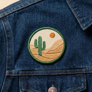 May include: Embroidered patch featuring a desert landscape with a green cactus, tan dunes, and a yellow sun against a cream background. The patch is circular with a green border and is affixed to a blue denim jacket.