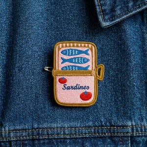 May include: A rectangular embroidered patch featuring a pink sardine can with three blue fish inside. The can has a gold border and a pull tab. Two red tomatoes are on the front, and the word "Sardines" is embroidered in white. A silver safety pin is attached.