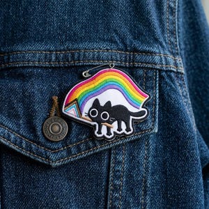 May include: A black cat patch with a rainbow and a transgender flag design, pinned to a denim jacket. The patch features a black cat silhouette with white details, a colorful rainbow, and a transgender flag. The jacket is a dark blue denim.