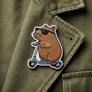 May include: A brown capybara wearing sunglasses rides a light blue scooter. The embroidered patch has a white border and is affixed to a green jacket. The capybara is the central focus of the patch.