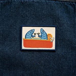 May include: Embroidered patch featuring two blue fish in a red sardine can, each holding a cocktail glass. The patch has a white background with a navy blue border, set against a dark blue denim backdrop.