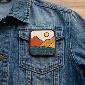 May include: Embroidered patch featuring a landscape design with mountains, trees, and a sun. The patch is square with rounded corners and a brown border. It is sewn onto a denim jacket pocket.