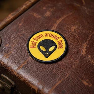 May include: A round patch with a yellow background and a black alien head design. The text "Not from around here" is in a circular pattern around the alien. The patch is on a brown leather surface.
