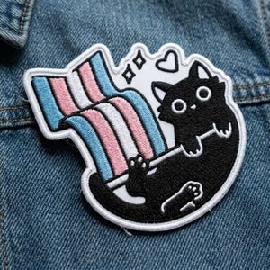 May include: Embroidered patch featuring a black cat with large eyes, paws, and a white outline. The cat is positioned beneath a rainbow-colored arc in the colors of the transgender flag. Two small diamonds and a heart are above the cat.