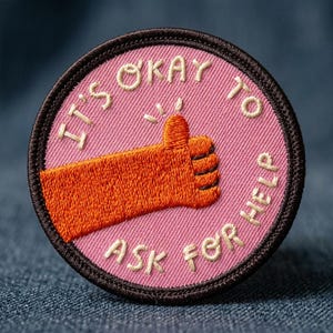 May include: Embroidered patch with a pink background and a brown border. The patch features an orange thumbs-up hand gesture and the text "IT'S OKAY TO ASK FOR HELP" in off-white thread. The patch is round and has a textured appearance.