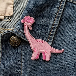 May include: Pink embroidered patch of a dinosaur wearing a cowboy hat, attached to a denim jacket. The dinosaur has a long neck and a small hat. The jacket has a metal button with a star design.