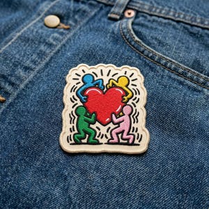 May include: A colorful patch featuring a red heart held by four stylized figures in blue, yellow, green, and pink. The patch has a cream-colored background with black outlines and radiating lines, and is sewn onto a denim fabric.
