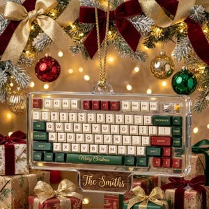 May include: A clear acrylic keyboard-shaped Christmas ornament featuring cream, dark green, and red keys. The space bar displays the text Merry Christmas in gold, and a wooden base at the bottom is engraved with the name The Smiths in gold script.