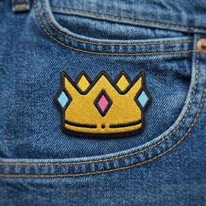 May include: Embroidered patch featuring a gold crown with black outlining. The crown has yellow, pink, and blue accents. The patch is placed on a blue denim pocket.
