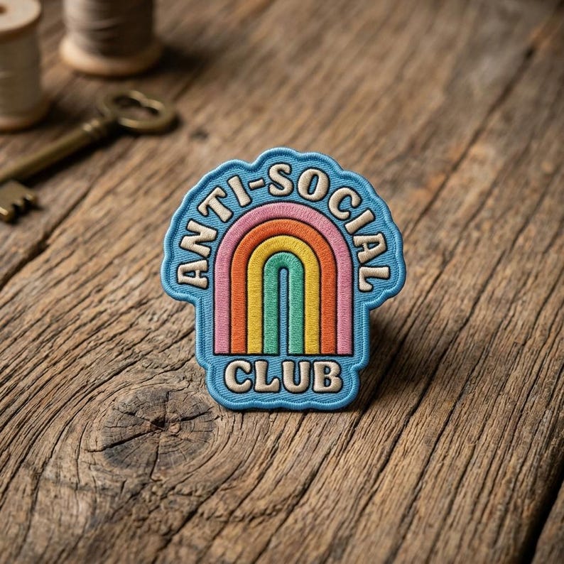 以下が含まれることがあります： 青いアーチ型の縁に「ANTI-SOCIAL CLUB」の文字が刺繍されたパッチ。パッチの中央には虹のデザインがあり、ピンク、オレンジ、黄色、緑などの色があります。パッチは木製の表面にあります。