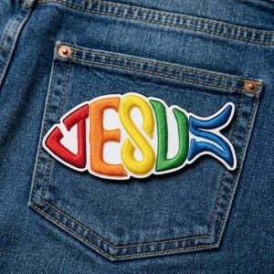 May include: A colorful patch in the shape of a fish, with the word "JESUS" spelled out in rainbow colors. The patch is sewn onto the back pocket of a pair of blue denim jeans.