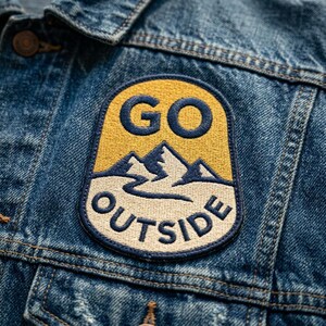 May include: Embroidered patch with the words "GO OUTSIDE" in navy blue on a yellow and off-white background. The patch features a mountain range design. The patch is sewn onto a blue denim jacket.