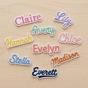 May include: A collection of embroidered name patches in various colors, including pink, purple, blue, yellow, and orange. Names like Claire, Lily, and Hannah are displayed in a cursive font with a white border. The patches are arranged on a light wood surface.