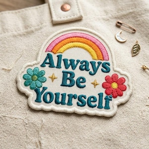 May include: Embroidered patch with the text "Always Be Yourself" in teal, featuring a rainbow, flowers, and stars. The patch is on a beige tote bag, with a crescent moon pin, leaf pin, and safety pins.