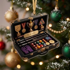 May include: A black briefcase-shaped Christmas ornament featuring a miniature pool set with billiard balls, cues, a rack, medals, a trophy, and purple gloves. A gold-colored plaque in the center displays the text NAME, suspended by a gold ribbon.