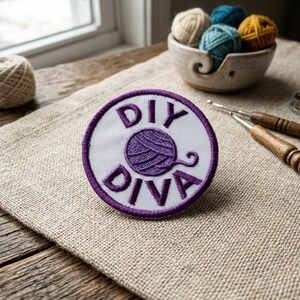 May include: A round white patch with purple text that reads "DIY DIVA" surrounds a purple yarn ball graphic. The patch is on a tan surface with a bowl of yarn and crochet hooks in the background. The patch is likely for a crafter.