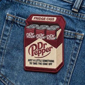 May include: Embroidered patch featuring a maroon box of Dr. Pepper cans labeled "Fridge Cigs." The patch includes the Dr. Pepper logo and the text "Just a little something to take the edge off." The patch is on a blue denim background.