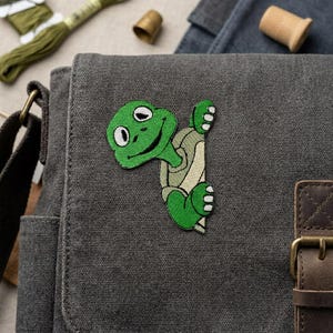 May include: A green embroidered turtle patch with a smiling face is affixed to a gray canvas messenger bag. The turtle has a light green shell and white accents. The bag has a brown leather strap and buckle.