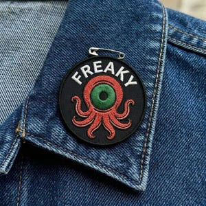 May include: A black circular patch with the word "FREAKY" in white lettering, featuring a red octopus-like design with a green eye. The patch is attached to a denim jacket with a silver safety pin.