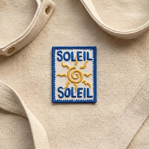 May include: A square patch with the word "SOLEIL" in blue, above and below a yellow sun design. The patch is on a beige canvas material, with a matching strap and metal button visible.