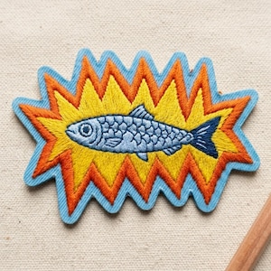 May include: Embroidered patch featuring a blue fish with detailed scales, set against a yellow and orange burst design, outlined in blue. The patch is likely for clothing or accessories.