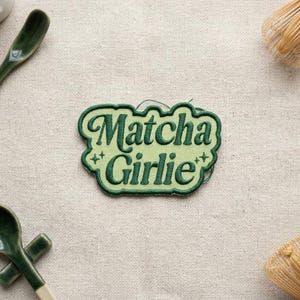 K&ouml;nnte beinhalten: Gr&uuml;ner, gestickter Aufn&auml;her mit dem Schriftzug "Matcha Girlie" in verspielter Schrift. Der Aufn&auml;her hat einen hellgr&uuml;nen Rand und ist mit kleinen Sternen verziert. Der Aufn&auml;her liegt auf einer beigen Leinenoberfl&auml;che.