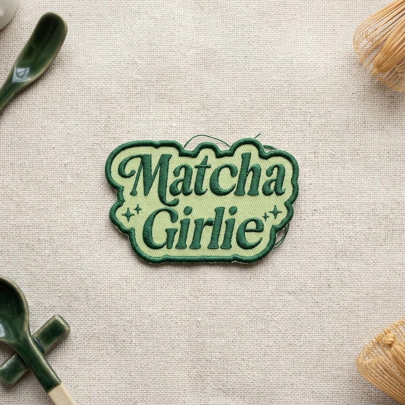 K&ouml;nnte beinhalten: Gr&uuml;ner, gestickter Aufn&auml;her mit dem Schriftzug "Matcha Girlie" in verspielter Schrift. Der Aufn&auml;her hat einen hellgr&uuml;nen Rand und ist mit kleinen Sternen verziert. Der Aufn&auml;her liegt auf einer beigen Leinenoberfl&auml;che.