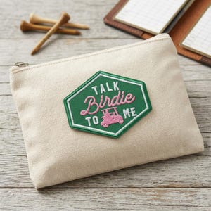 May include: A beige canvas zippered pouch with a green and pink embroidered patch that reads "Talk Birdie To Me" with a golf cart graphic. Wooden golf tees are in the background.
