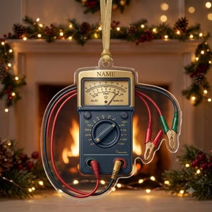 May include: A flat, clear acrylic ornament shaped like a multimeter with a gold-toned top section labeled NAME, a central analog gauge with star icons, a rotary dial, and two sets of red and black test leads with alligator clips attached.