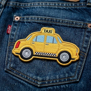 May include: Yellow embroidered taxi patch with black and white checkered accents, blue windows, and the word "TAXI" on top. The patch is sewn onto a blue denim pocket.