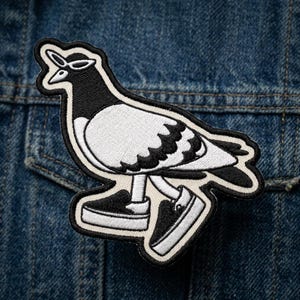 May include: Embroidered patch featuring a stylized pigeon wearing sunglasses and sneakers. The patch is primarily black and white with a cream-colored border. The pigeon is depicted walking, with its feet in black and white sneakers. The patch is placed on a denim jacket.