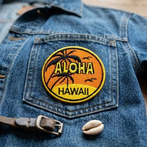 May include: Close-up of a denim jacket with a circular patch. The patch features the words "Aloha Hawaii" with palm trees and birds in orange and black. A brown leather belt and a seashell are also visible.