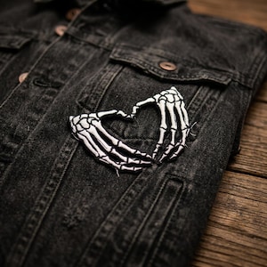 May include: A black denim jacket with a white and black skeleton hand heart patch. The patch is embroidered and sewn onto the jacket's chest. The jacket has two chest pockets and copper-colored buttons.