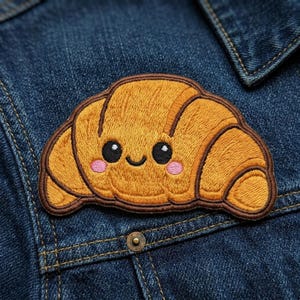 May include: Embroidered patch of a smiling croissant with a cute face. The patch features shades of brown and orange, with pink cheeks and black eyes. The patch is sewn onto a blue denim jacket.