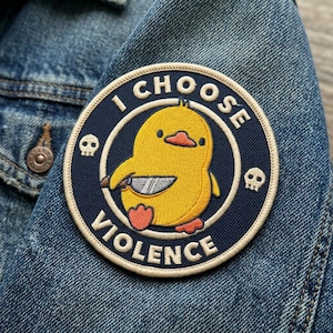 May include: A round patch with the text "I CHOOSE VIOLENCE" encircling a yellow cartoon duck holding a knife. The patch has a navy blue background with two skull icons and is sewn onto a denim jacket.