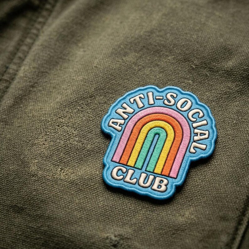 以下が含まれることがあります： 「ANTI-SOCIAL CLUB」の白い文字と虹のデザインが特徴的なカラフルなパッチ。パッチは水色の縁取りがあり、質感のあるオリーブグリーンの生地に配置されています。