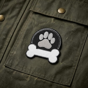 May include: Close-up of a dark olive green jacket pocket with a circular patch. The patch features a black background with a white bone and a gray paw print. The jacket has two brass-colored buttons.