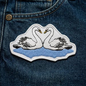 May include: Embroidered patch featuring two white swans with black detailing, forming a heart shape with their necks. The swans are set against a light blue water design, all on a white background. The patch is sewn onto a blue denim fabric.