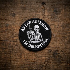May include: A black circular patch with white text and a skeleton illustration. The text reads "AS FAR AS I KNOW I'M DELIGHTFUL". The skeleton is holding a cup of coffee. The patch is on a wooden surface.