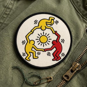 May include: A round patch featuring a Keith Haring-style design of three figures in yellow and red, holding hands around a yellow sun. The patch has a black border and is set against a green fabric background.