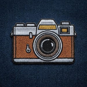 May include: Embroidered patch of a vintage camera with a brown body, silver top, and black lens. The camera design features a detailed lens, buttons, and a flash mount. The patch is outlined in a silver border and is set against a dark blue denim background.