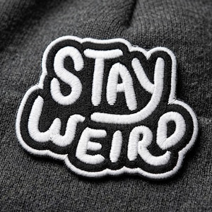 May include: A dark gray beanie with a black and white embroidered patch. The patch features the words "STAY WEIRD" in a bold, stylized font. The beanie is made of a soft, knitted material.