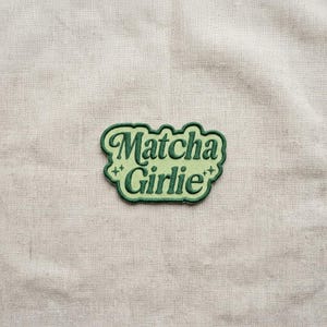 K&ouml;nnte beinhalten: Ein gr&uuml;n-hellgr&uuml;ner, gestickter Aufn&auml;her mit dem Schriftzug "Matcha Girlie" in einer verspielten Schriftart. Der Aufn&auml;her hat einen hellgr&uuml;nen Hintergrund und einen dunkelgr&uuml;nen Rand. Auf beiden Seiten des Textes befinden sich zwei kleine sternf&ouml;rmige Designs.